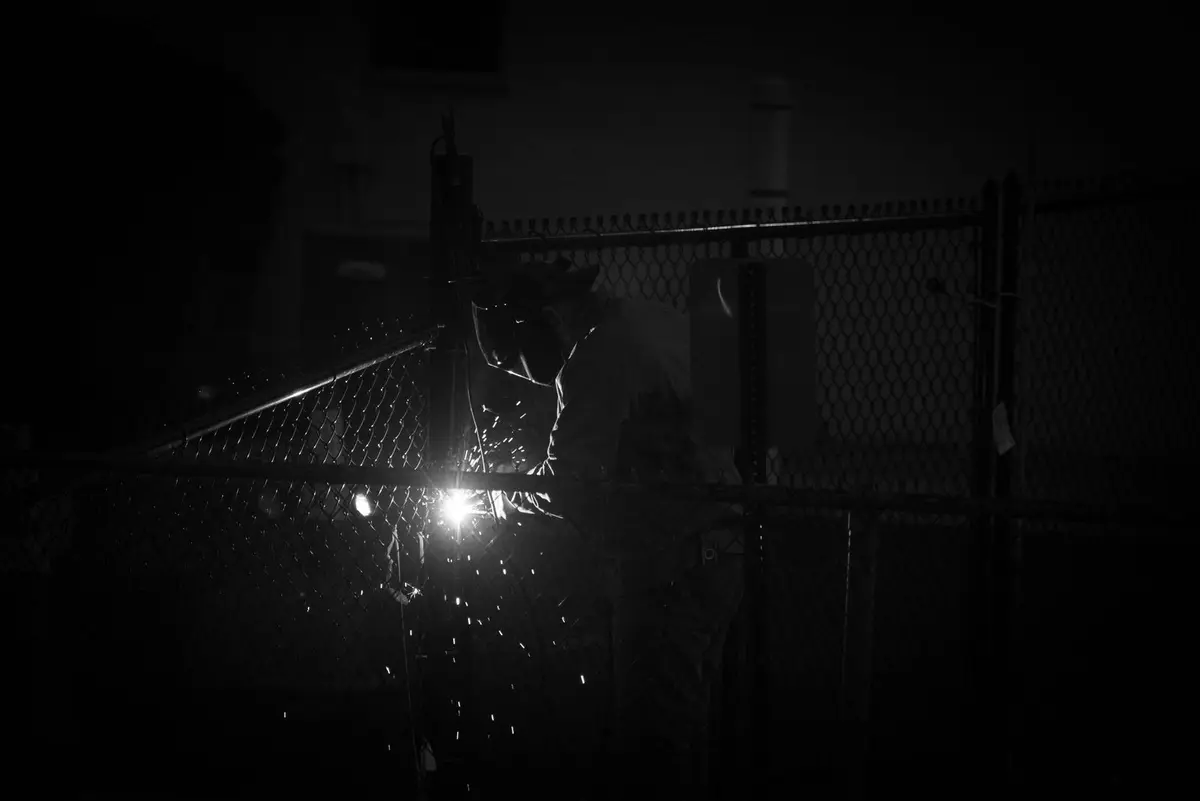 Welder in a helmet welding a chain-link fence gate post at night, sparks glowing